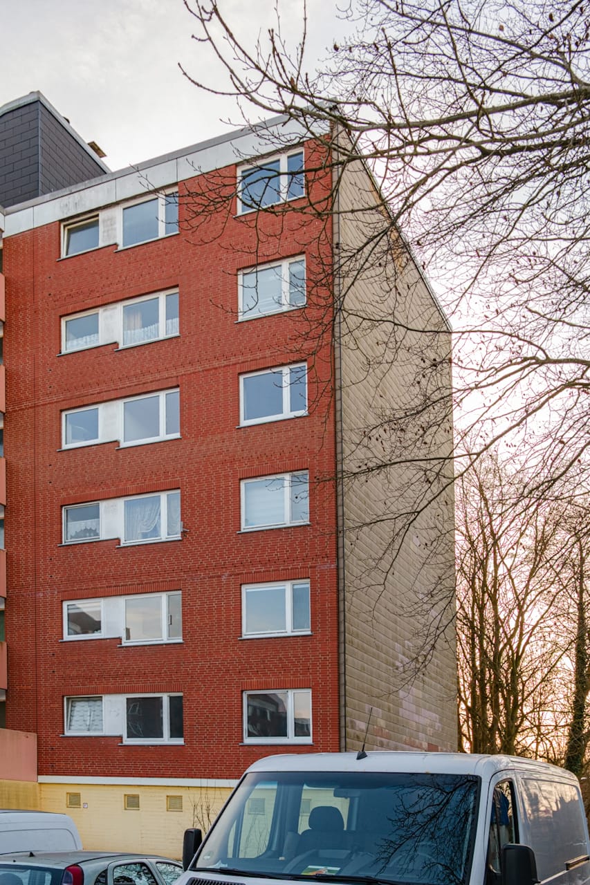Stirnseite eines roten Mehrfamilienhauses mit heller Schindelverkleidung an der Giebelseite. Die ungewöhnliche Gebäudeform verweist auf einen nie fertiggestellten Bauabschnitt.
