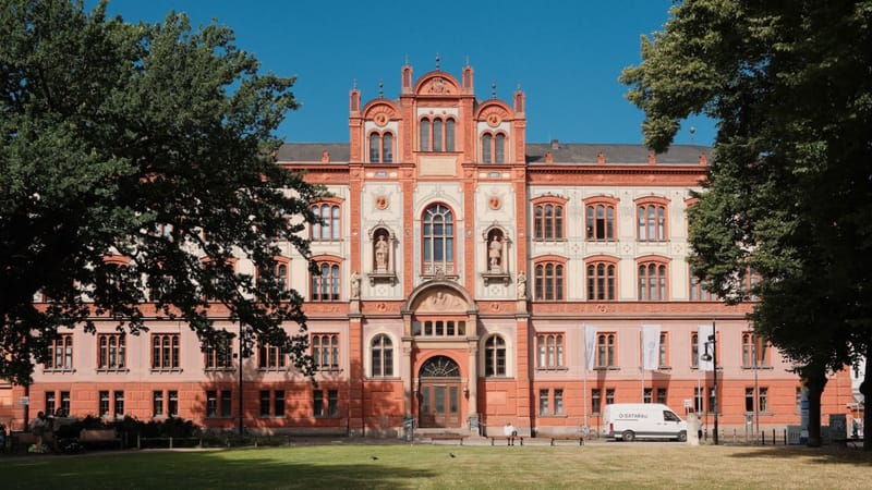 Das Universitäts-Hauptgebäude Rostock im warmen Sommerlicht, umgeben von Bäumen und blauem Himmel – ein Beispiel für die weiche norddeutsche Lichtstimmung.